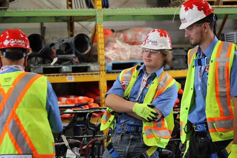 Two trainees in hard hats, safety glasses and vests standing beside warehouse shelving